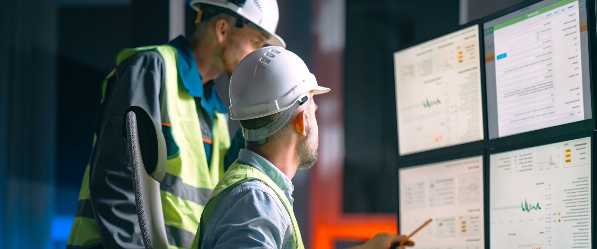Two individuals wearing hard hats and safety vests are in a control room. They are focused on multiple monitors displaying graphs and data, suggesting an industrial or technical setting.