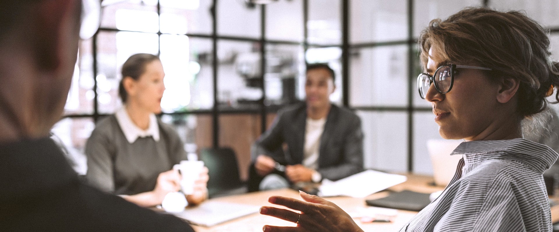 Four coworkers in a modern office meeting, one person speaking while others listen around a table with a laptop and documents