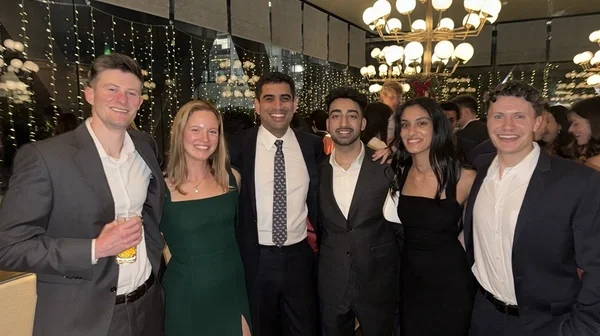 It shows a group of six people dressed in formal attire, smiling and standing together at an indoor event. The background features string lights and a decorative chandelier.