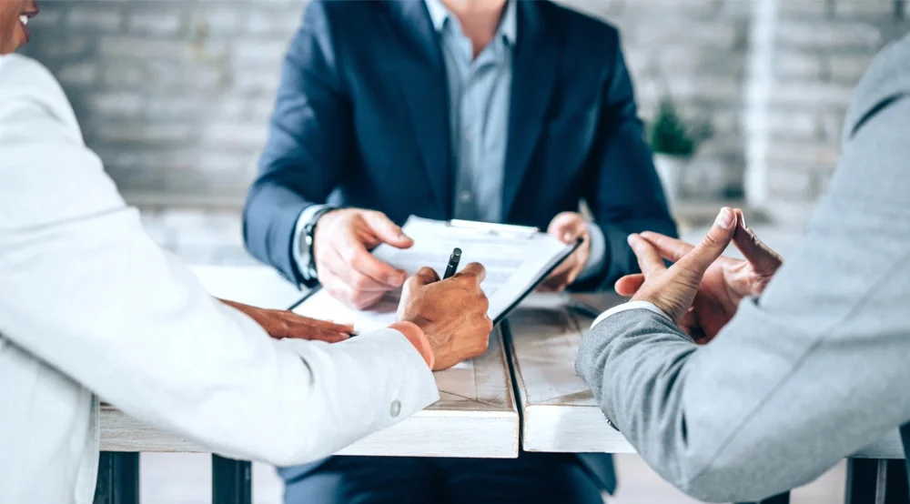 Three individuals are seated at a table in a business meeting. One person holds a clipboard with documents, while the others engage in discussion. The setting is professional, with a blurred background.