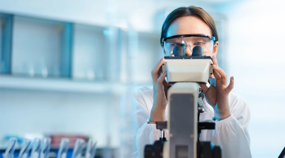 A person wearing safety goggles is using a microscope in a laboratory setting. Shelves with laboratory equipment are blurred in the background.